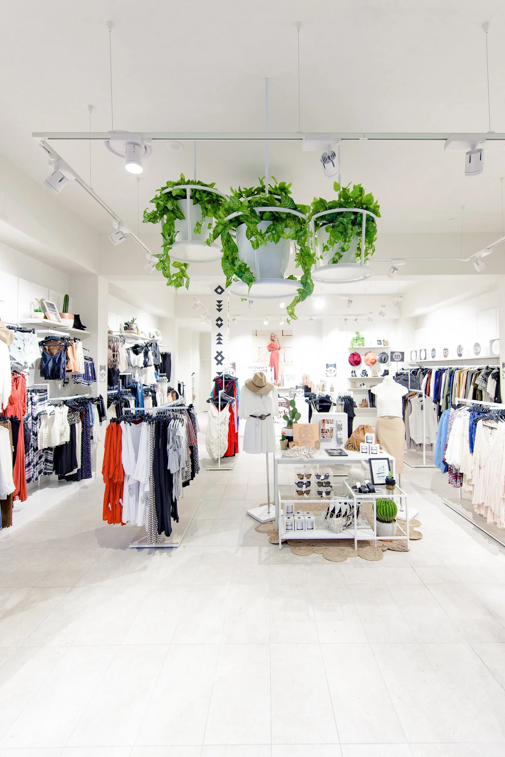Interior of a clothing boutique with hanging racks and shelves filled with clothes, accessories, and decorative items. Green plants hang from the ceiling at the center.