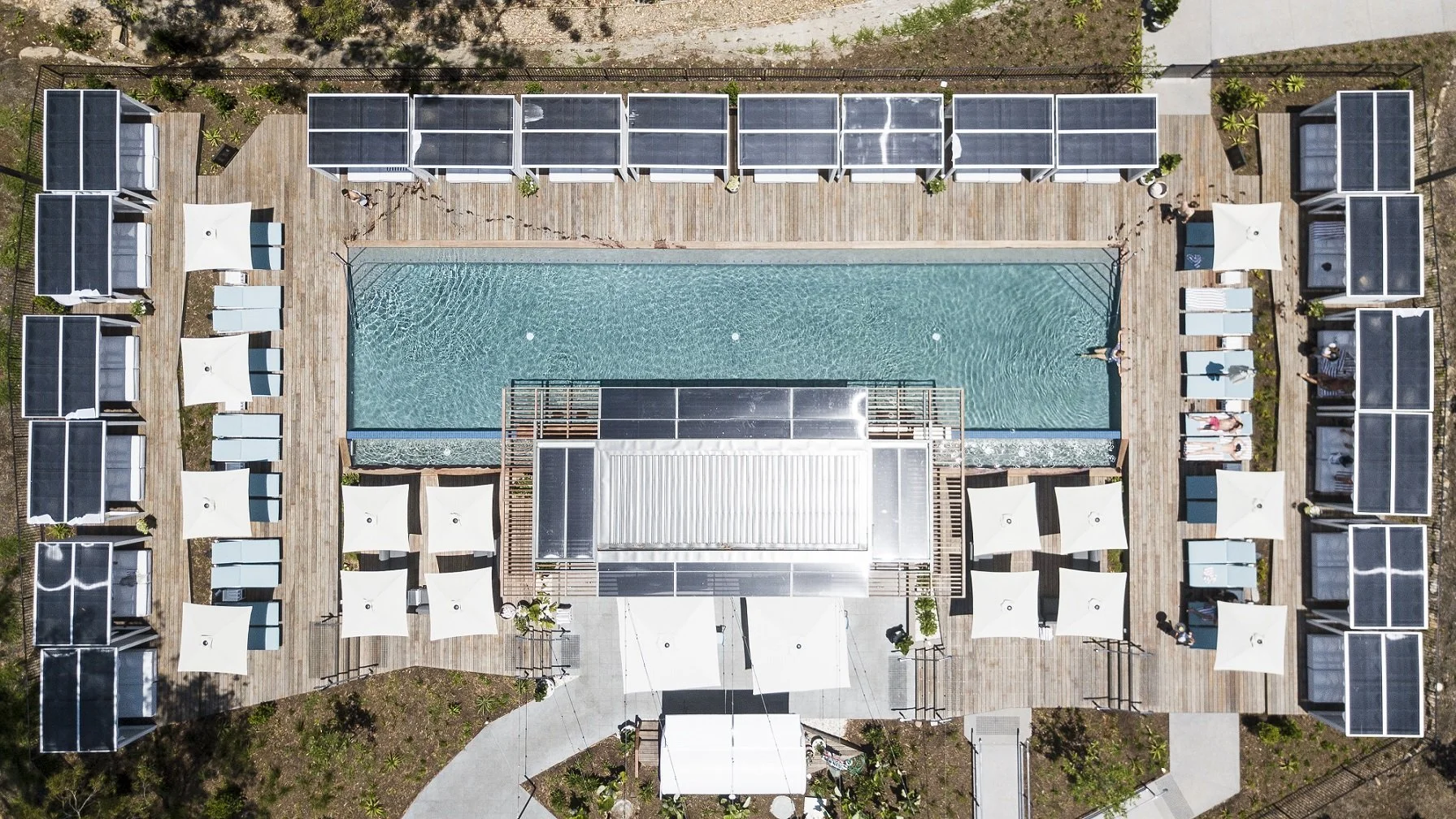 Aerial view of a swimming pool surrounded by cabanas and lounge chairs with umbrellas, with a wooden deck and landscaping.