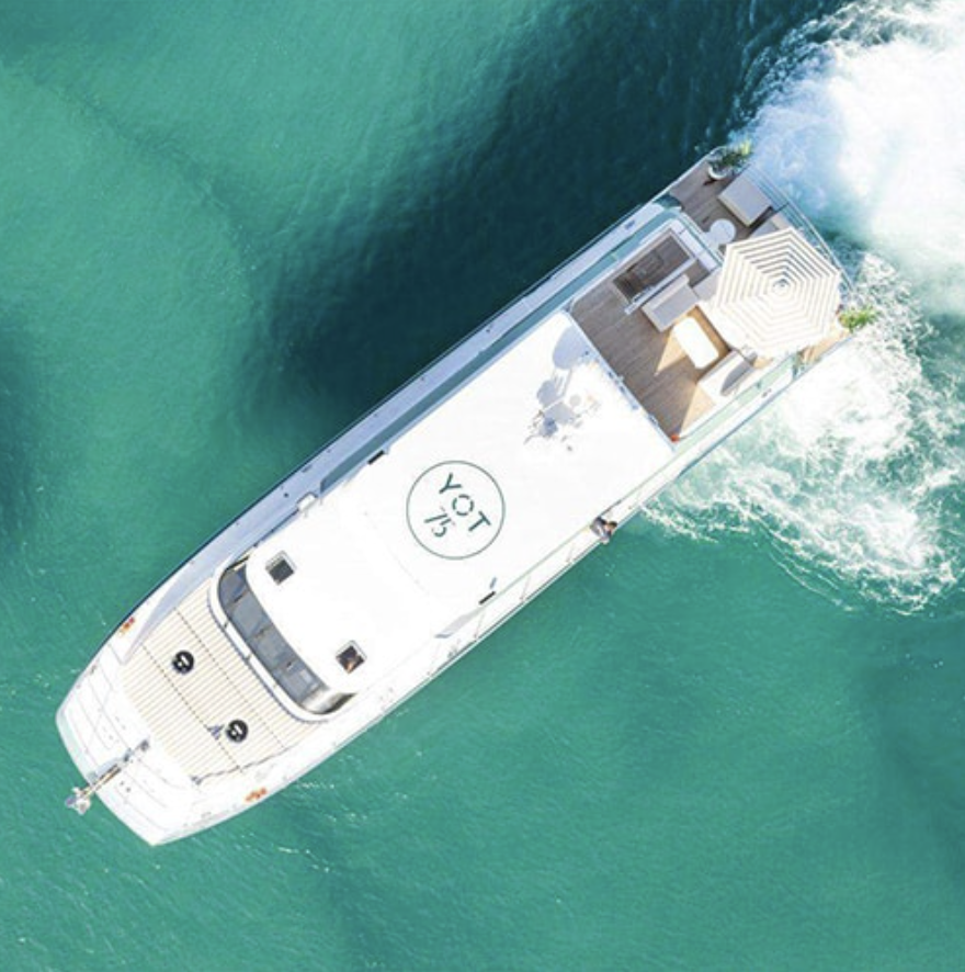 An aerial view of a white yacht cruising in turquoise water, with a rooftop deck featuring an umbrella, lounge chairs, and seating area.