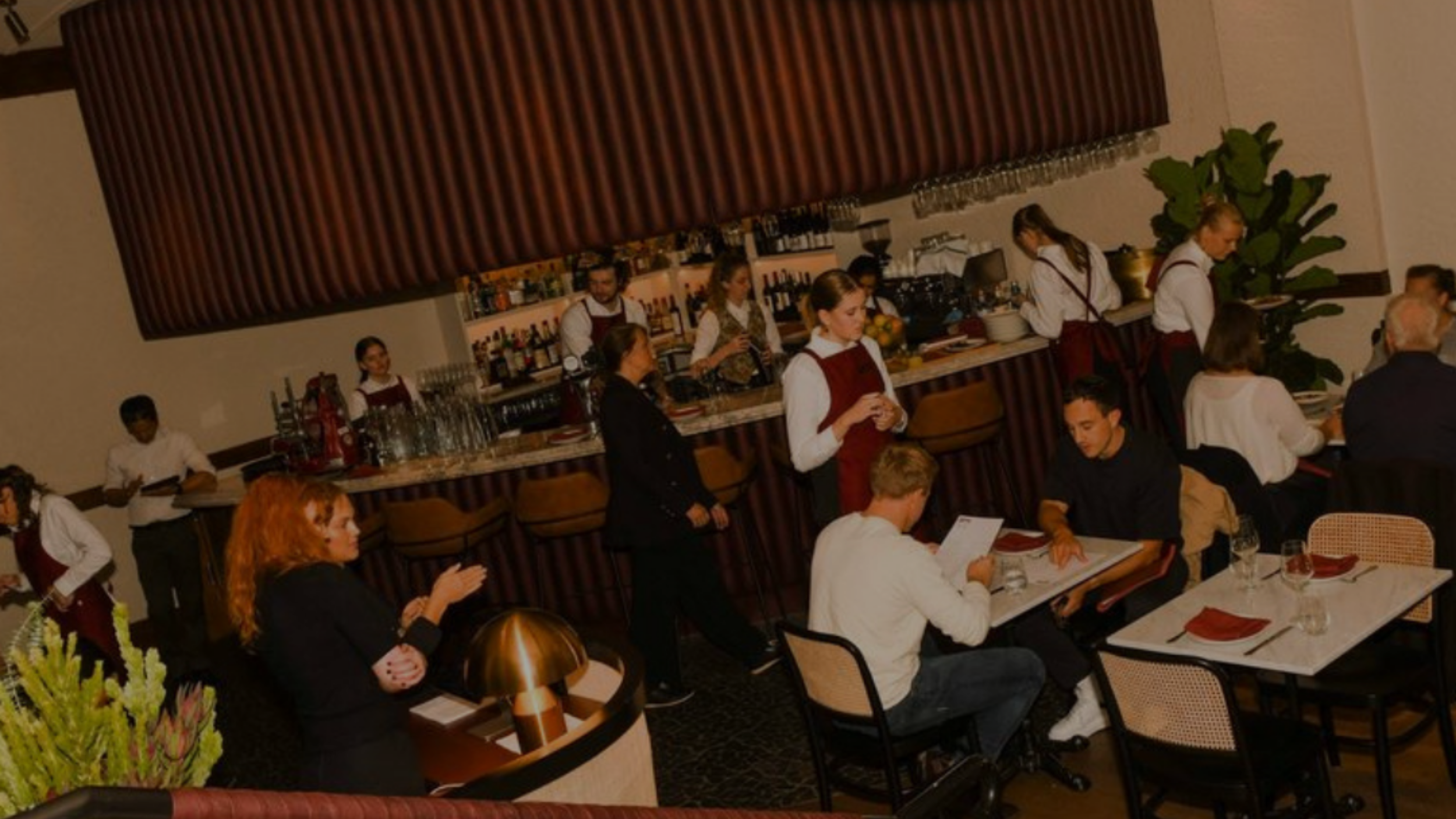 Restaurant scene with waitstaff in uniform, customers seated at tables, and a bar area in the background.