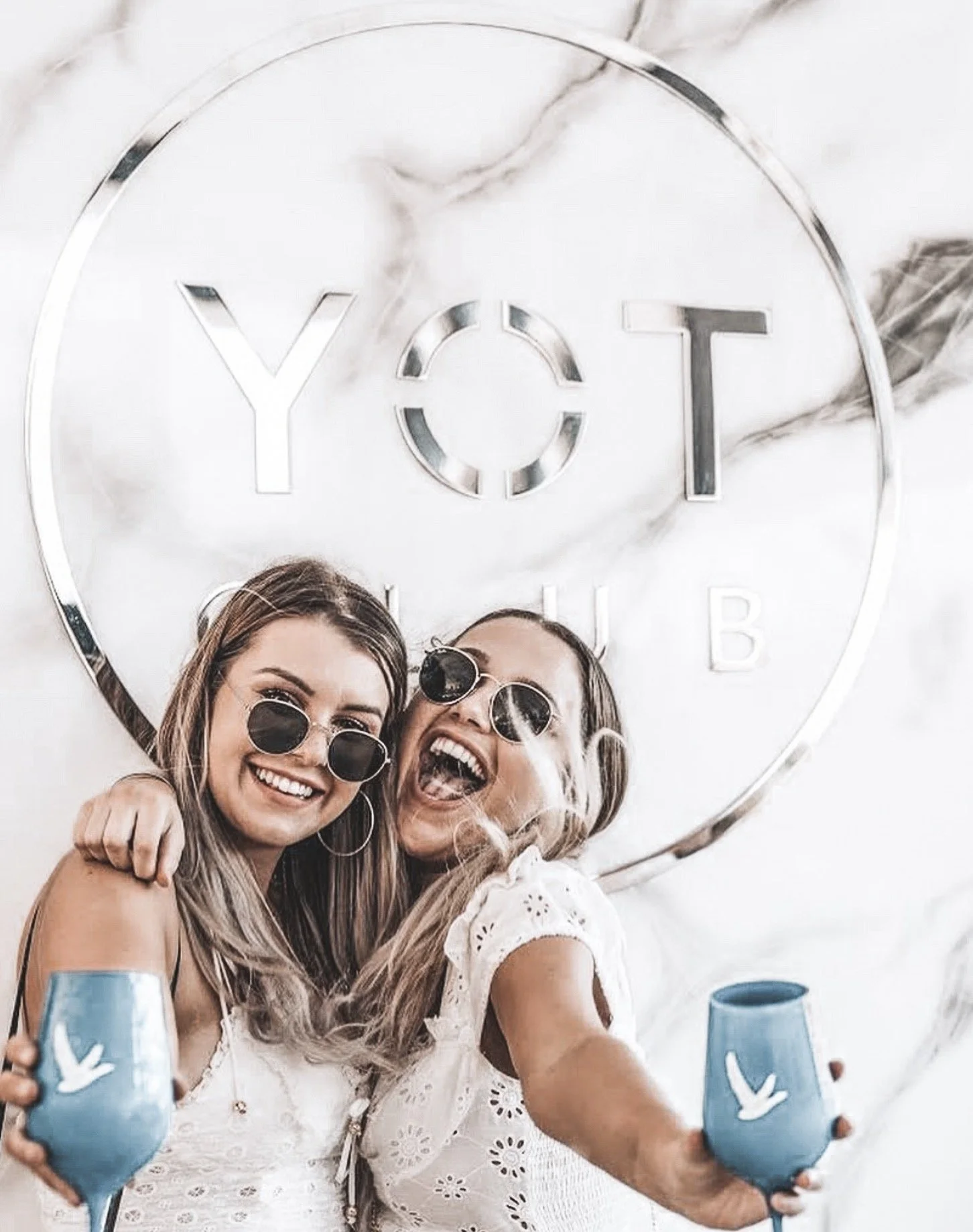 Two women smiling and celebrating, holding blue cups with white dove symbols, in front of a sign that reads 'YOT CLUB' on a marble wall.