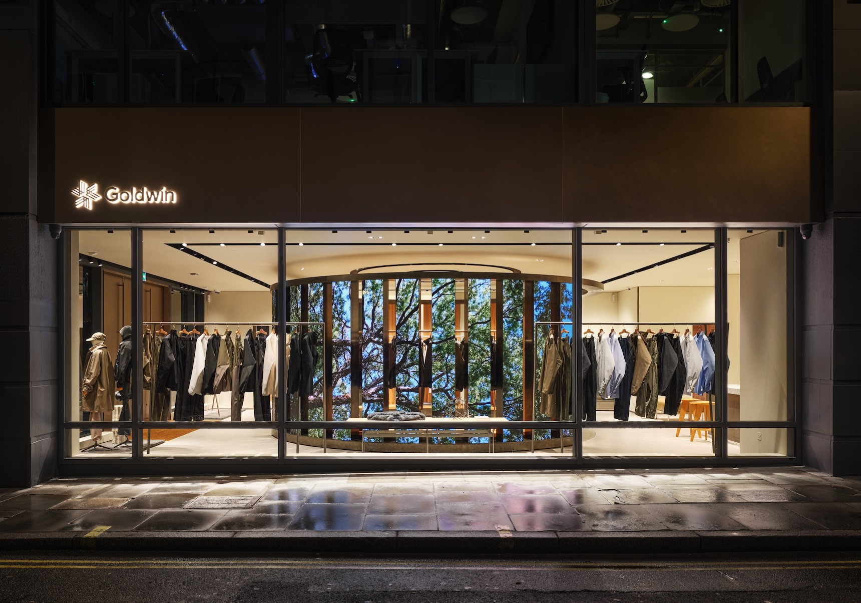 Goldwin’s glazed London storefront at night, with timber columns and a ring of illuminated digital screens visible inside.