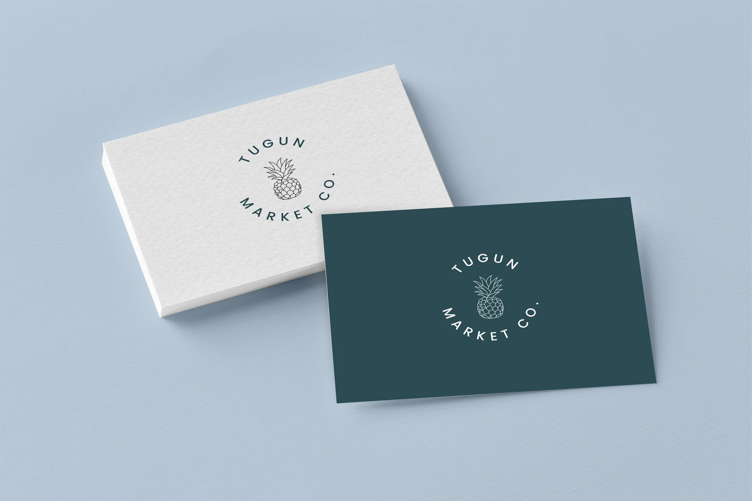 Two business cards for Tugun Market Co., one white and one dark teal, with a pineapple logo in the center, on a light blue surface.