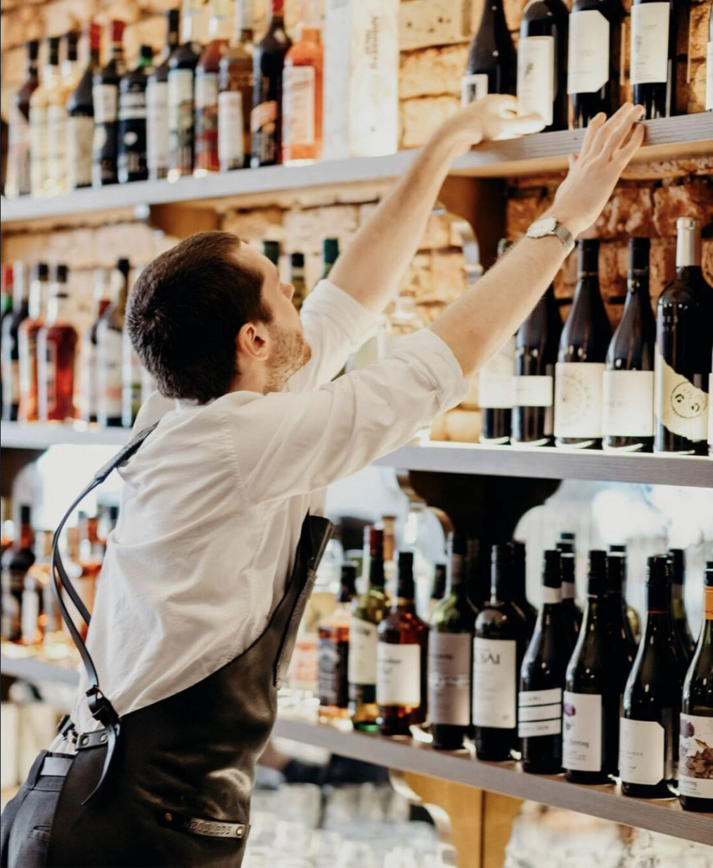 A man in a white shirt and black apron reaching for a bottle on a shelf filled with wine bottles in a wine shop or liquor store.