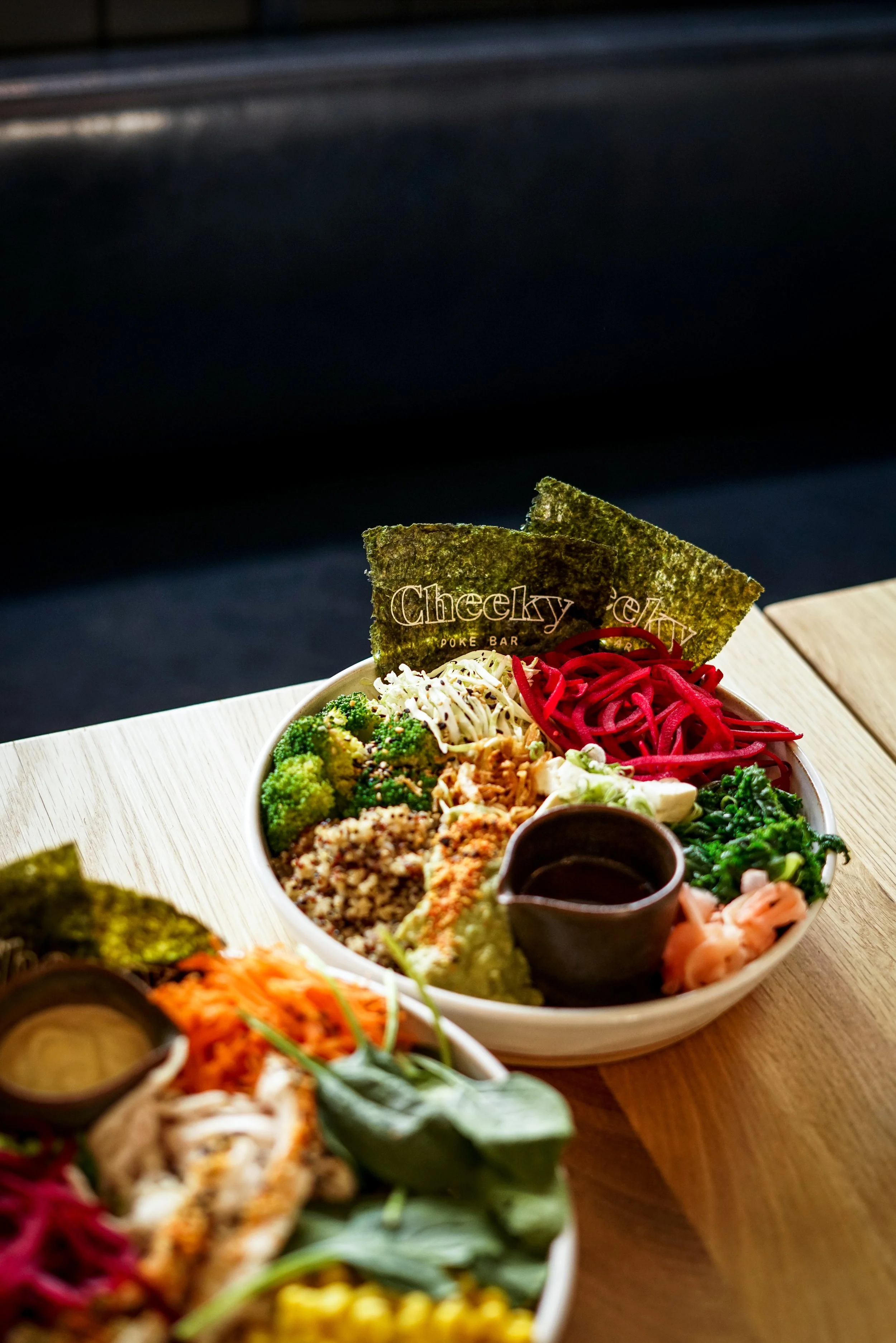 A bowl of assorted poke ingredients including vegetables, fish, and seaweed, with a small sauce container, on a wooden table at a poke bar.