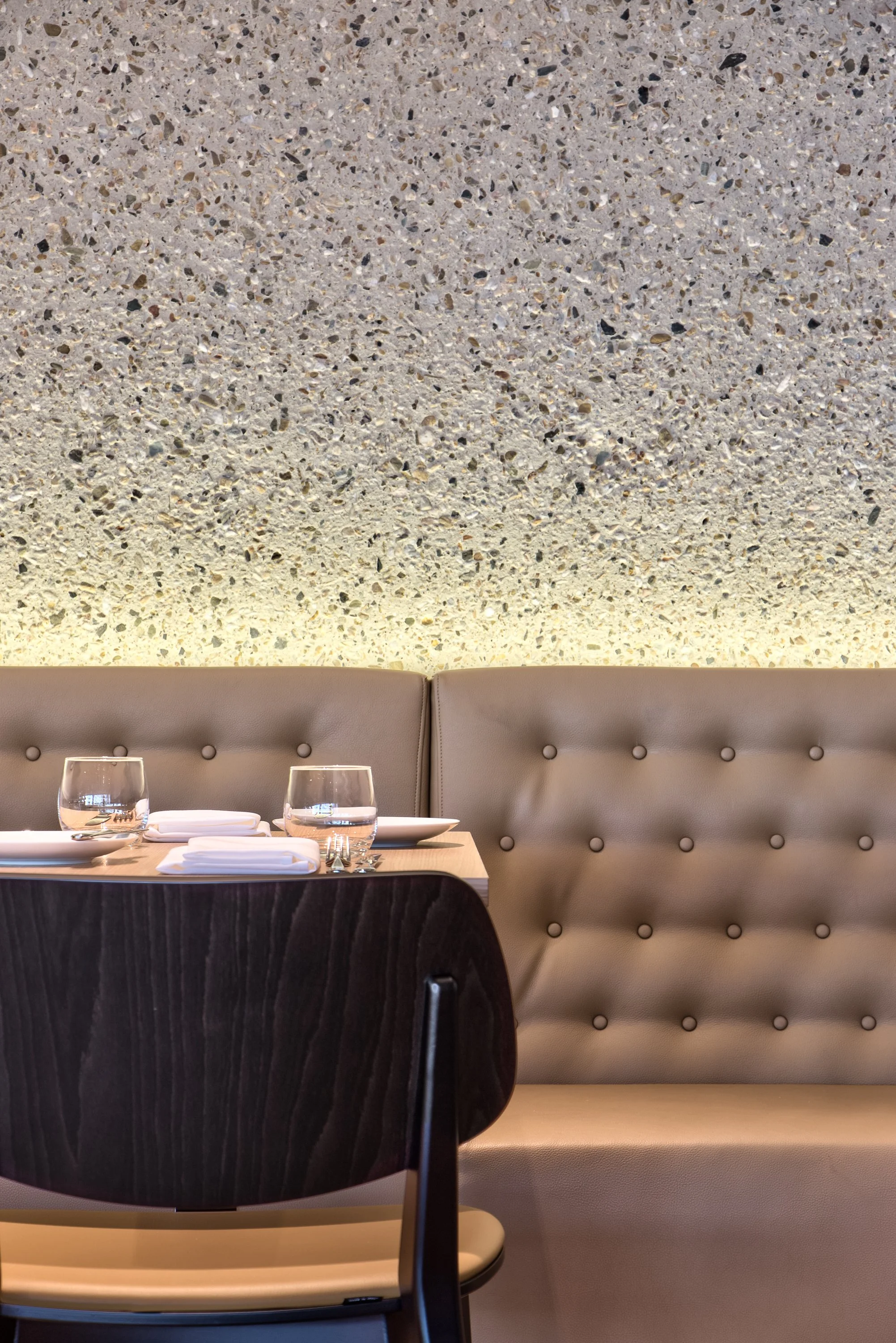 A restaurant table set with glasses, plates, and napkins, situated in front of a beige tufted bench against a speckled textured wall.