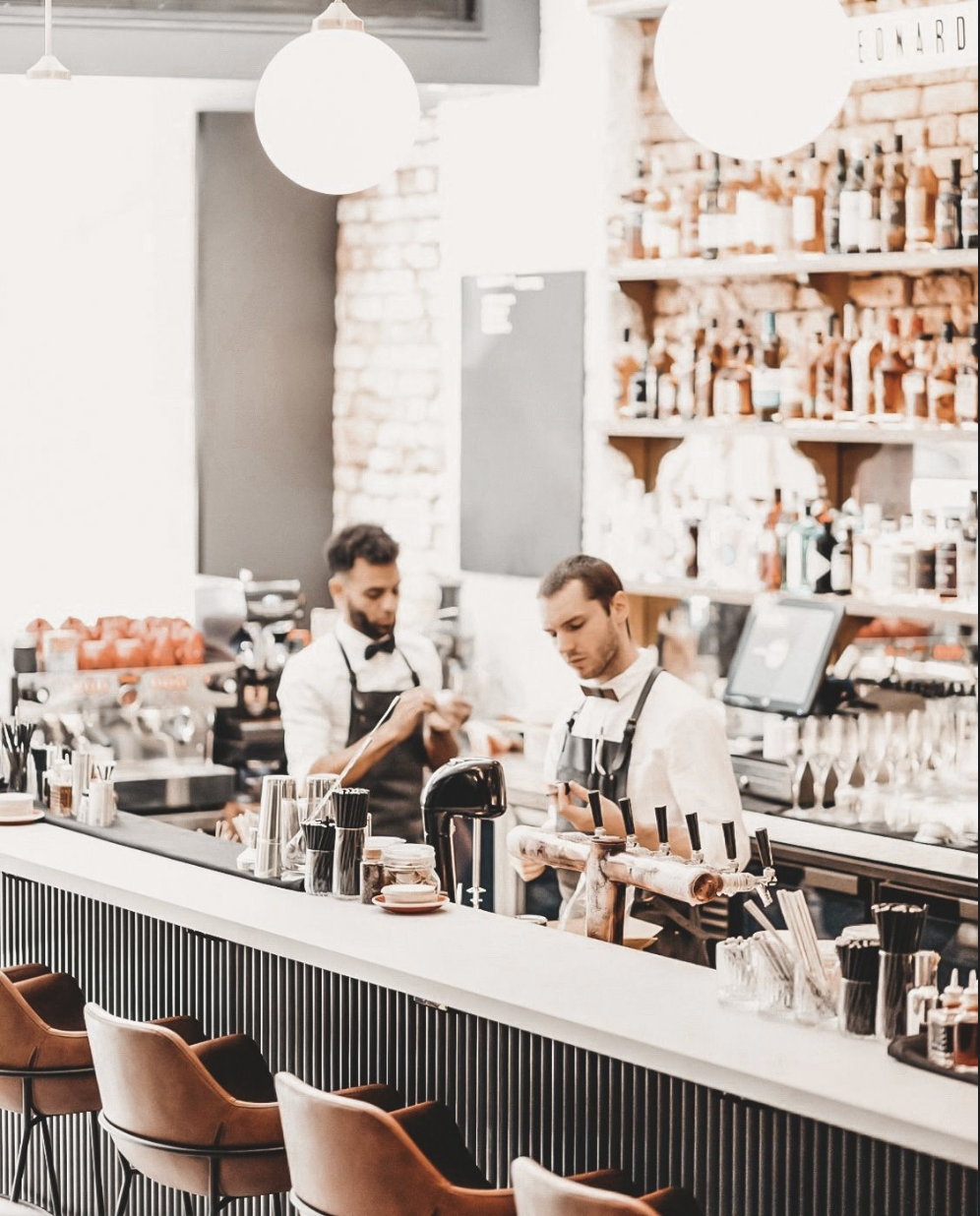 Baristas working behind a bar in a modern cafe or bar with brick and white walls, shelves with bottles, and brown chairs in the foreground.