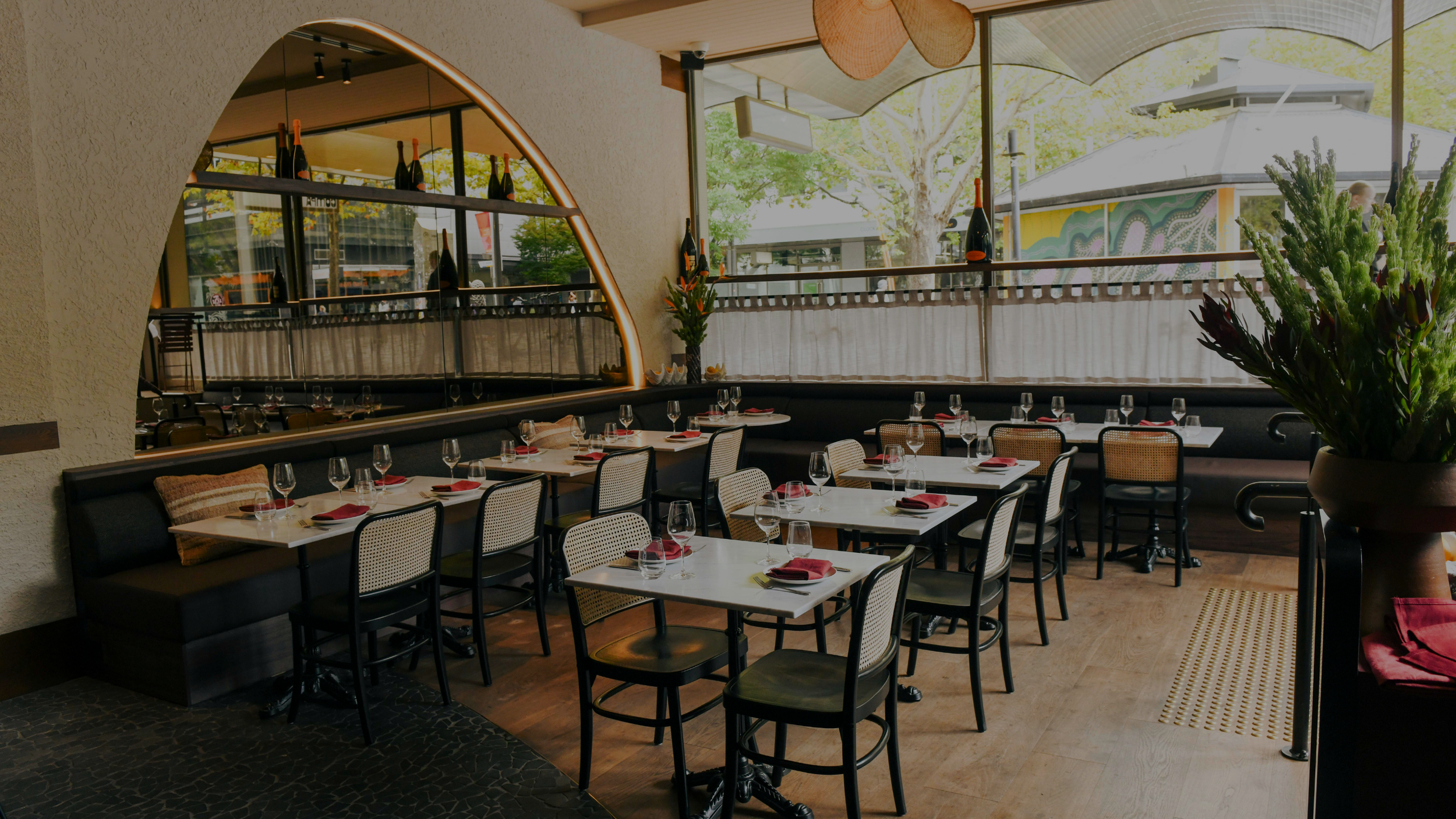Empty restaurant dining area with neatly set tables, wine glasses, red napkins, and wicker chairs, large window with trees outside, and modern decor.