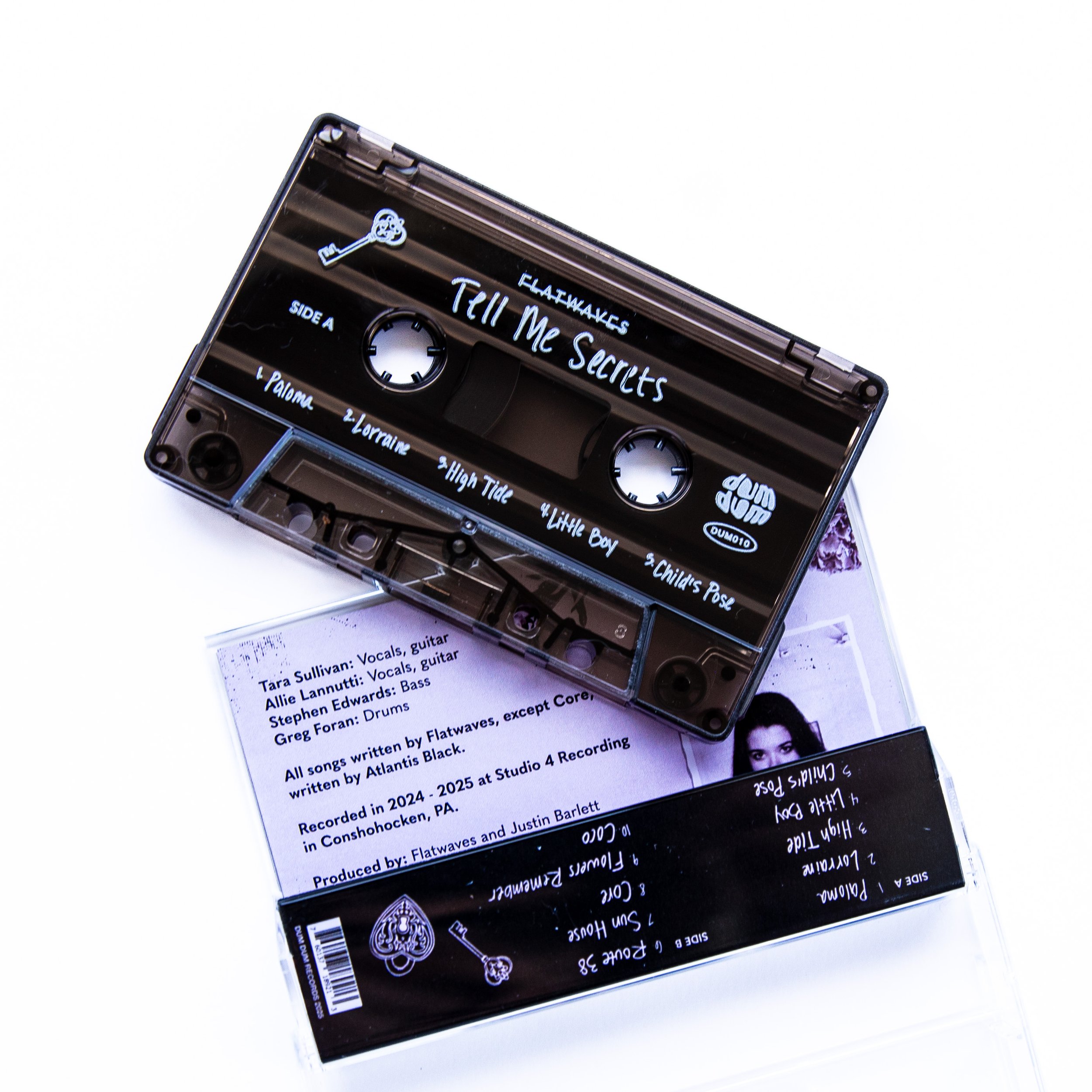 A black cassette tape titled "Tell Me Secrets" by Flatwaves resting on a piece of paper with song credits and a booklet with photos inside.