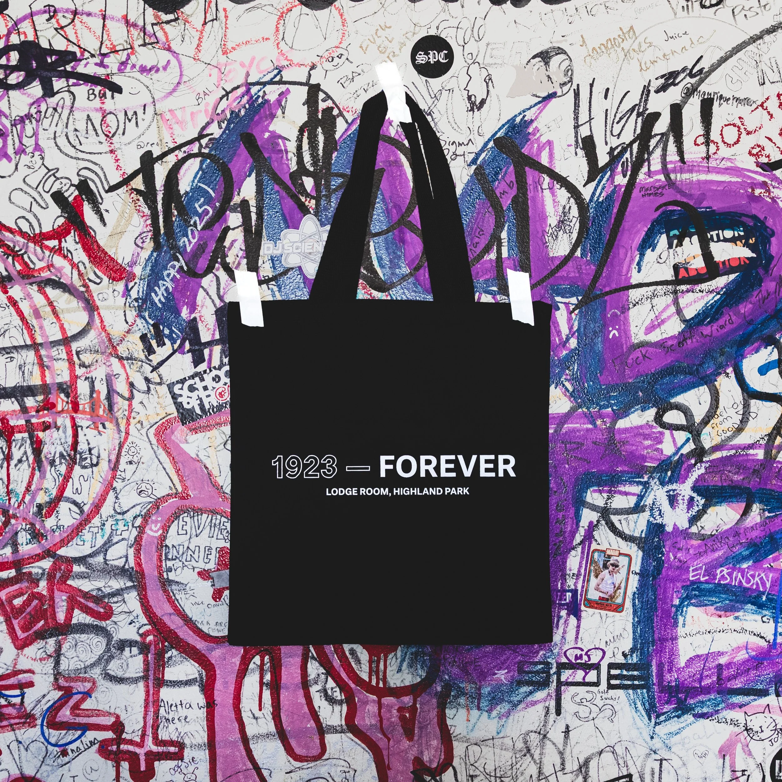 Black tote bag with the text "1923 — FOREVER" and "LODGE ROOM, HIGHLAND PARK" on it, hanging against a graffiti-covered wall.