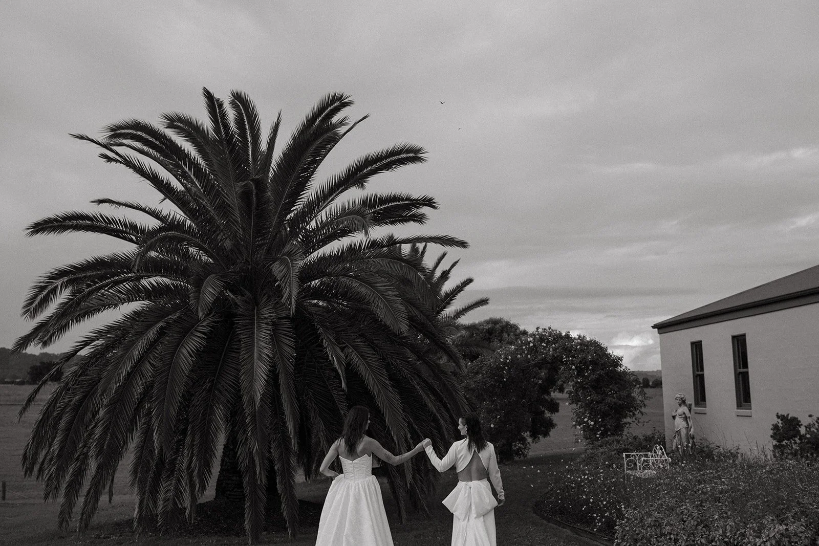 Two women in white dresses holding hands under a large palm tree outside near a building and garden, overcast sky.