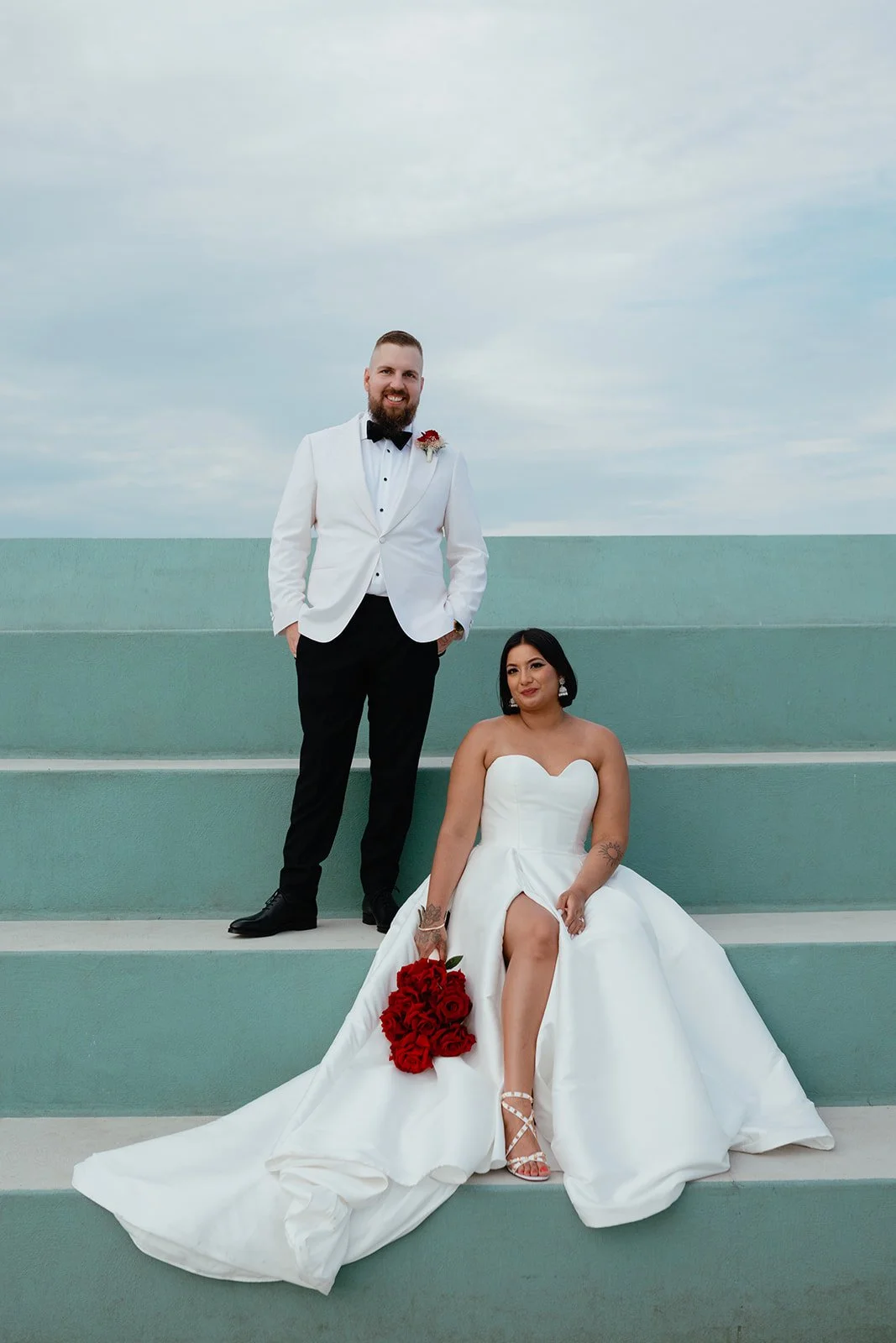A bride in a strapless white wedding gown sitting on steps, holding a bouquet of red roses, and a groom in a white tuxedo jacket with black pants standing beside her on the steps, both smiling outdoors against a sky background.