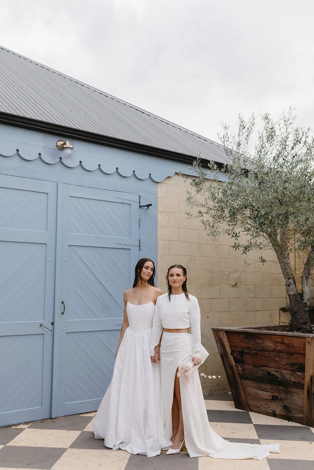 Two women wearing white dresses holding hands stand outdoors, near a blue barn door and a large planter with a tree, on a checkered pavement.