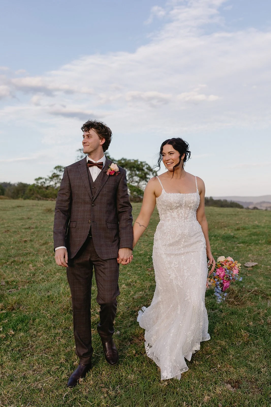 A couple dressed in wedding attire walking hand in hand in an open grassy field with trees and a blue sky with clouds in the background. The bride is holding a colorful bouquet and is smiling, while the groom is looking to the side.