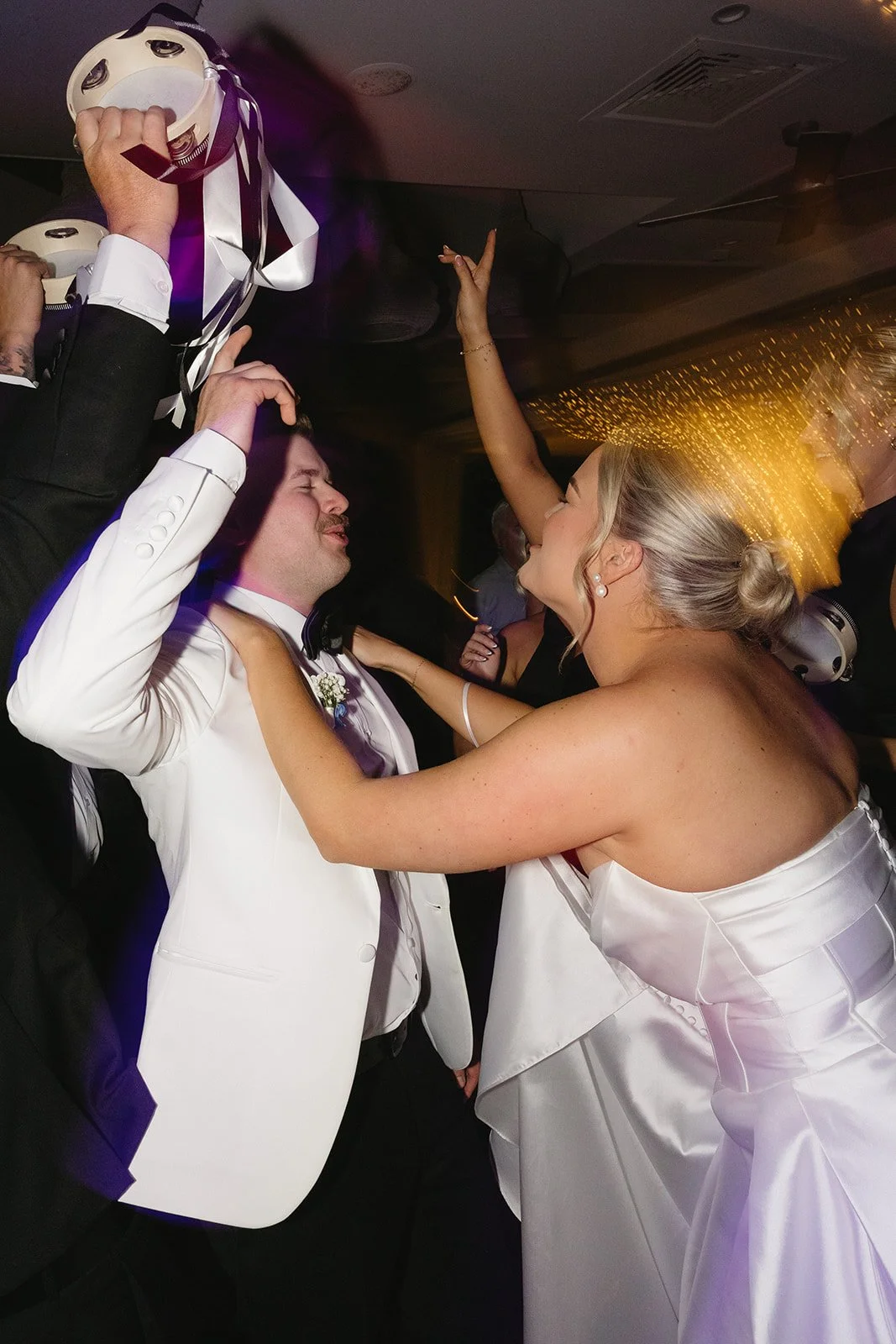 A bride and groom dancing closely at their wedding reception, with the bride wearing a white strapless dress and the groom in a white tuxedo jacket, surrounded by guests celebrating.