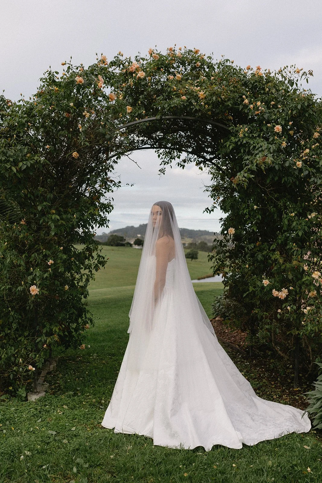 A woman in a white wedding dress and veil standing under a floral arch outdoors, with a grassy field and hills in the background.