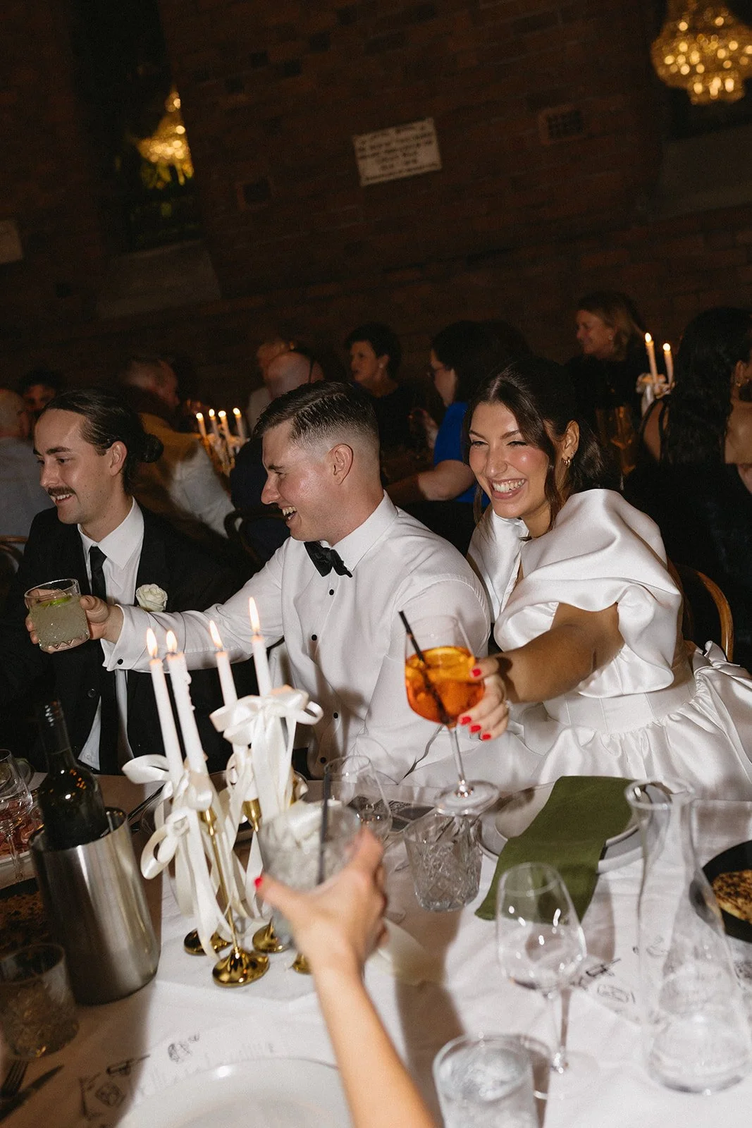 A group of people at a wedding reception, sitting at a table with candles, drinks, and celebratory decorations, smiling and toasting.