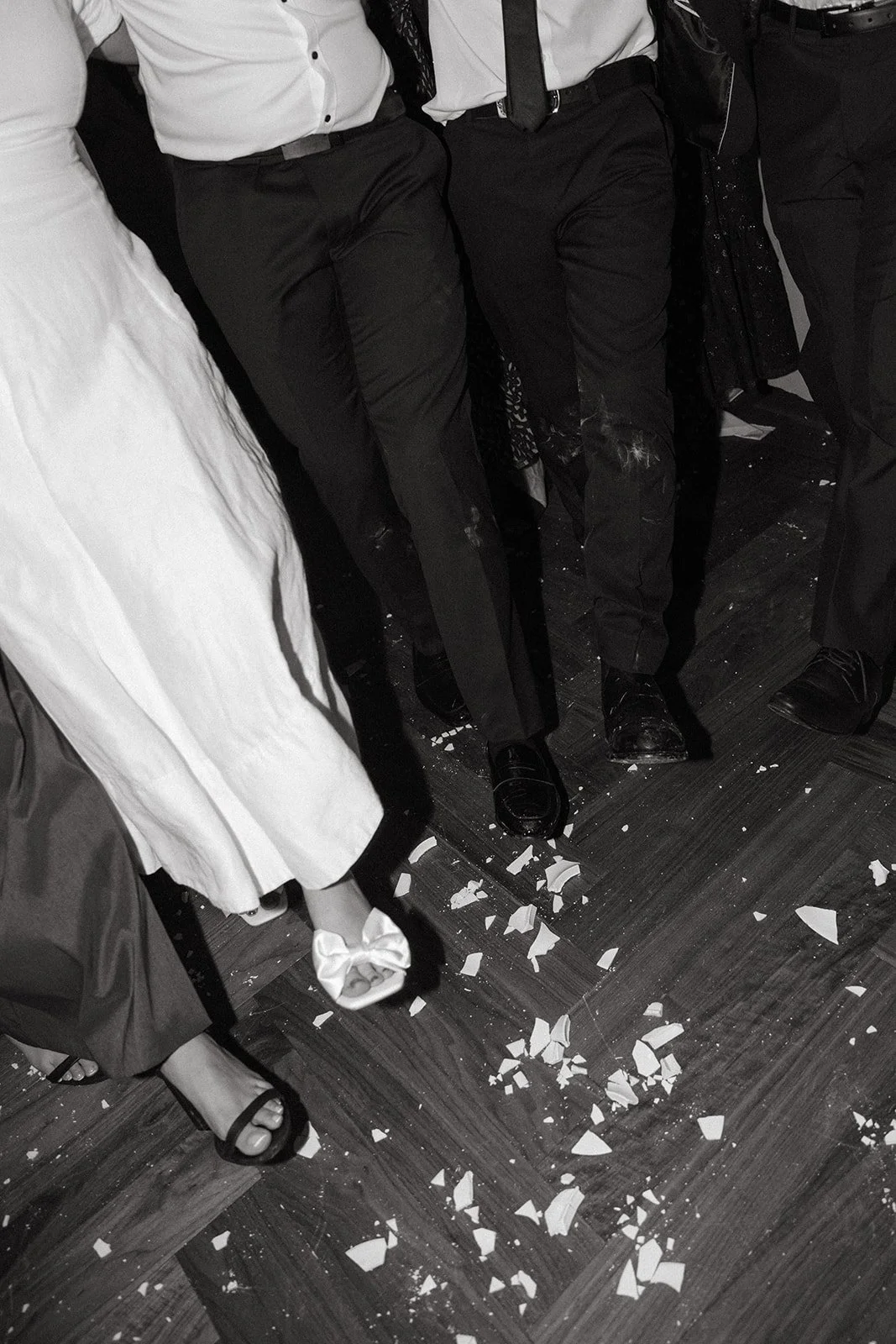 People sitting on chairs, with a woman in a white dress and black sandals, and several men in suits with black shoes, with broken pieces of white ceramic or porcelain on the floor.