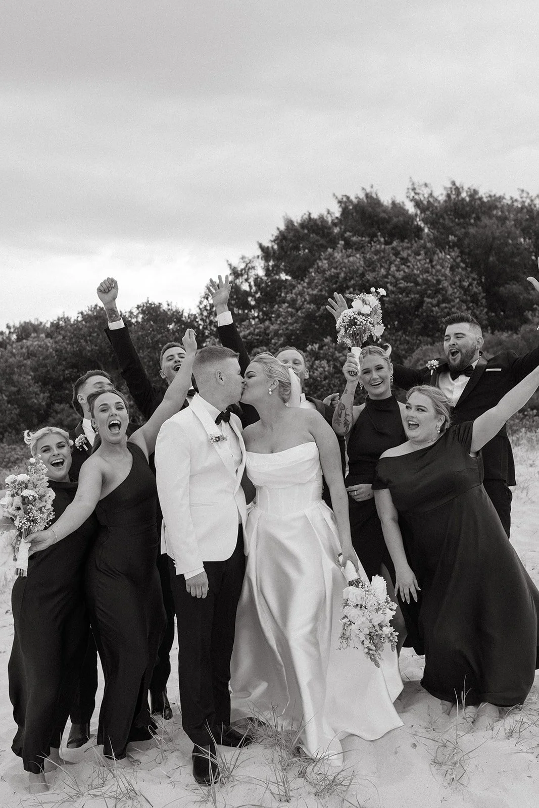 Black and white photo of a wedding party on a beach, with the bride and groom kissing, surrounded by bridesmaids and groomsmen celebrating with raised hands and smiling.