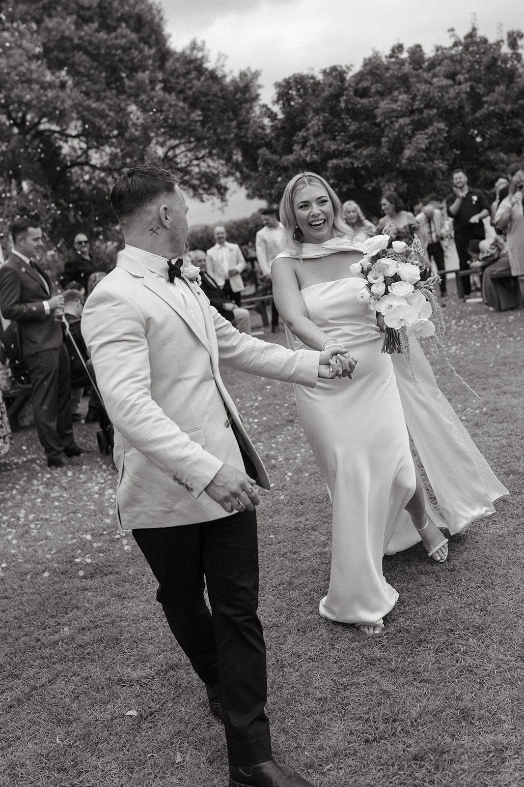 A black and white photo of a wedding celebration outdoors with a couple holding hands and smiling, surrounded by friends and family.