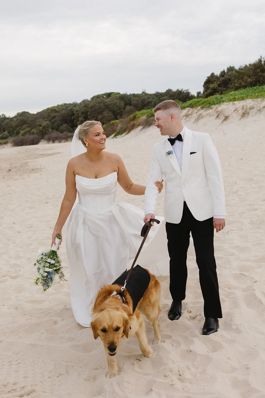 Bride and groom walking on the beach with dog, bride holding bouquet, groom in tuxedo with bow tie, bride in strapless wedding dress and veil, holding a leash, beach with sand dunes and greenery in background.