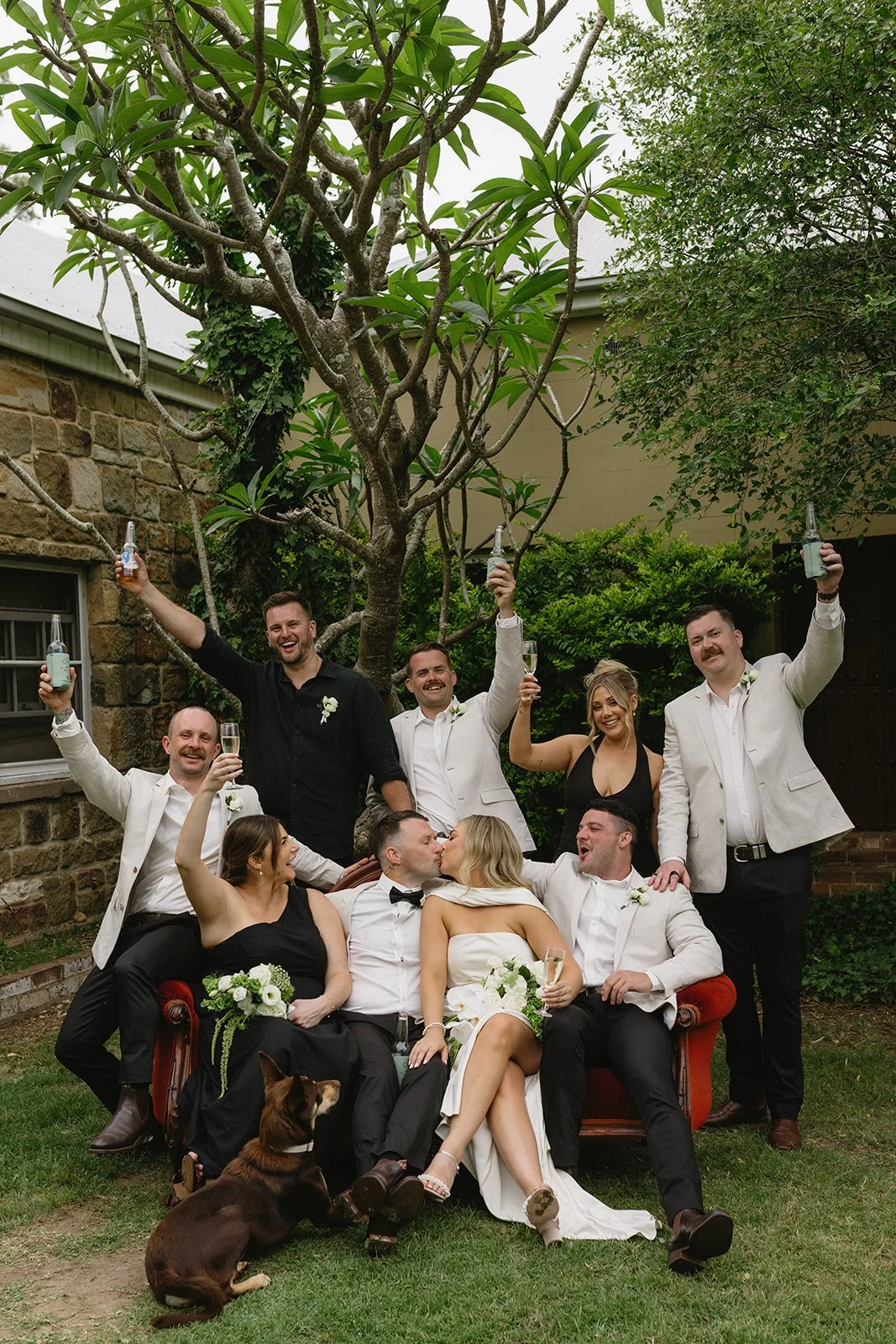 Wedding party celebrating outdoors, the bride and groom sitting on a vintage red sofa, kissing, surrounded by friends raising drinks, with a dog sitting on the grass in front, under a tree.