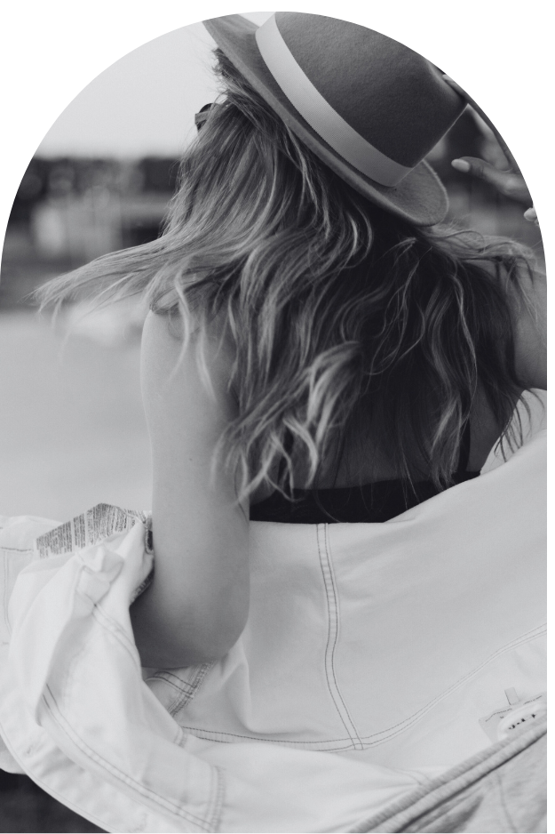 Een vrouw, gezien van achteren, met een hoed en een jasje die los over haar ellebogen hangt.