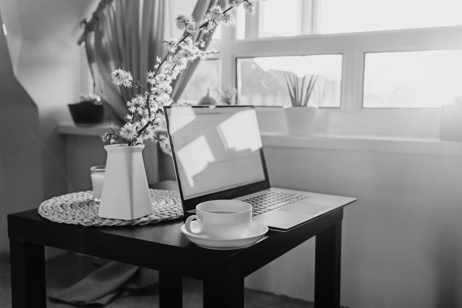 Zwart-witte foto van een tafel bij het raam met een open laptop en een kop thee, met bloemen op de achtergrond.