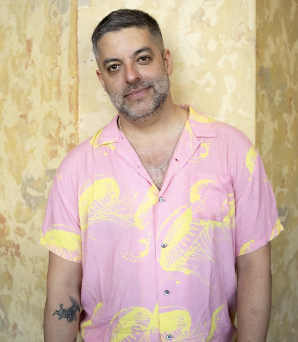  A light-skinned masculine person lighting smiling into the camera wearing a pink and yellow shirt against a mustard background 