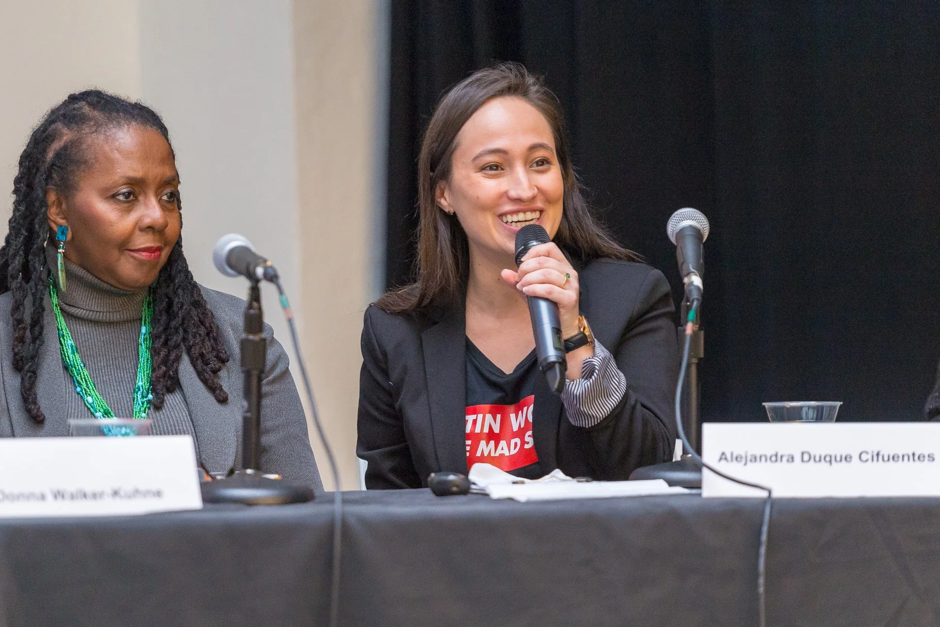 Alejandra is speaking at an event, holding a cordless microphone. She is wearing a black blazer and is smiling as she speaks to the crowd.