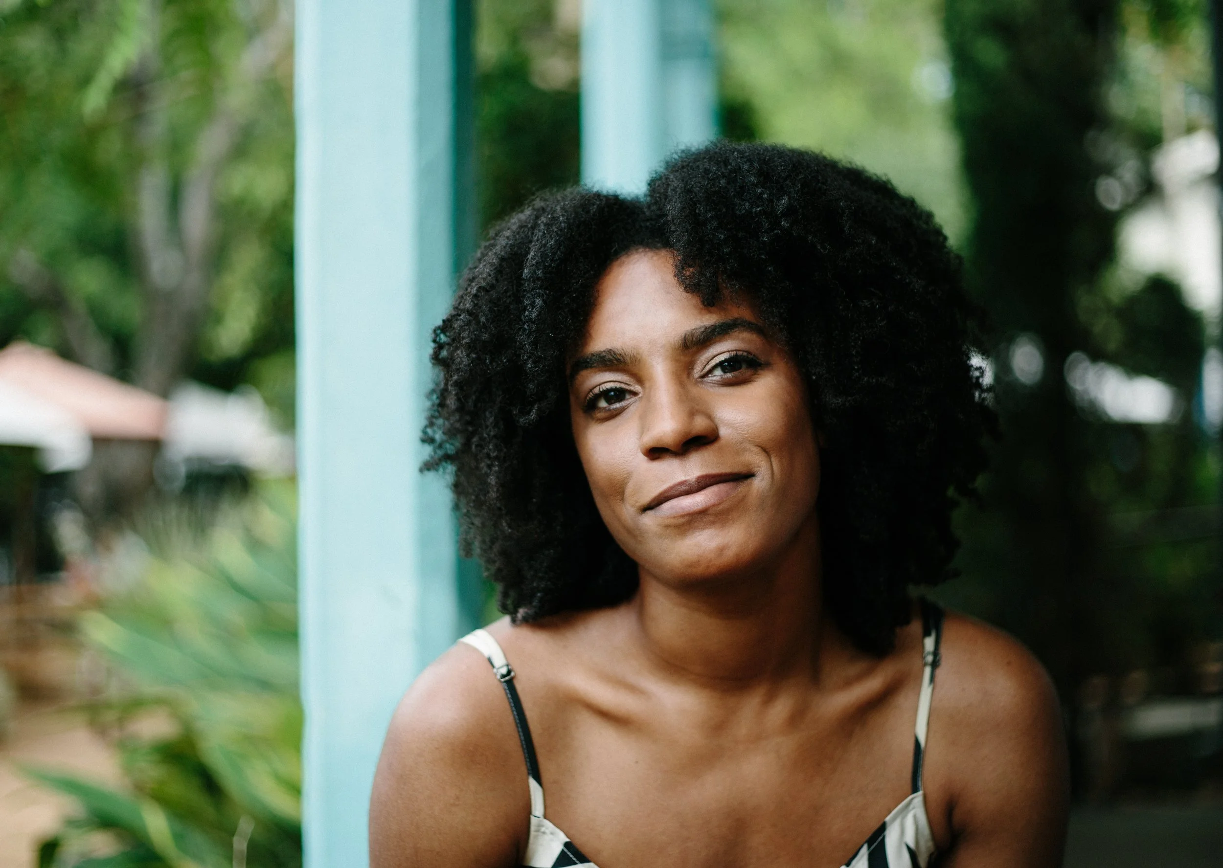 A black woman with black natural afro faces the camera, smirking. She's wearing a silk, houndstooth camisole.