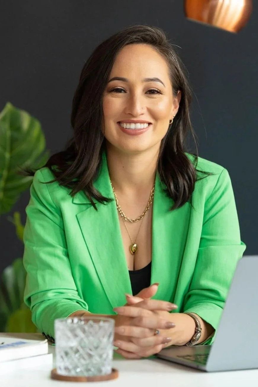 A photo of Alejandra Duque Cifuentes sat at a desk and wearing a light green blazer in a confident pose with clasped hands. She smiles warmly at the camera.