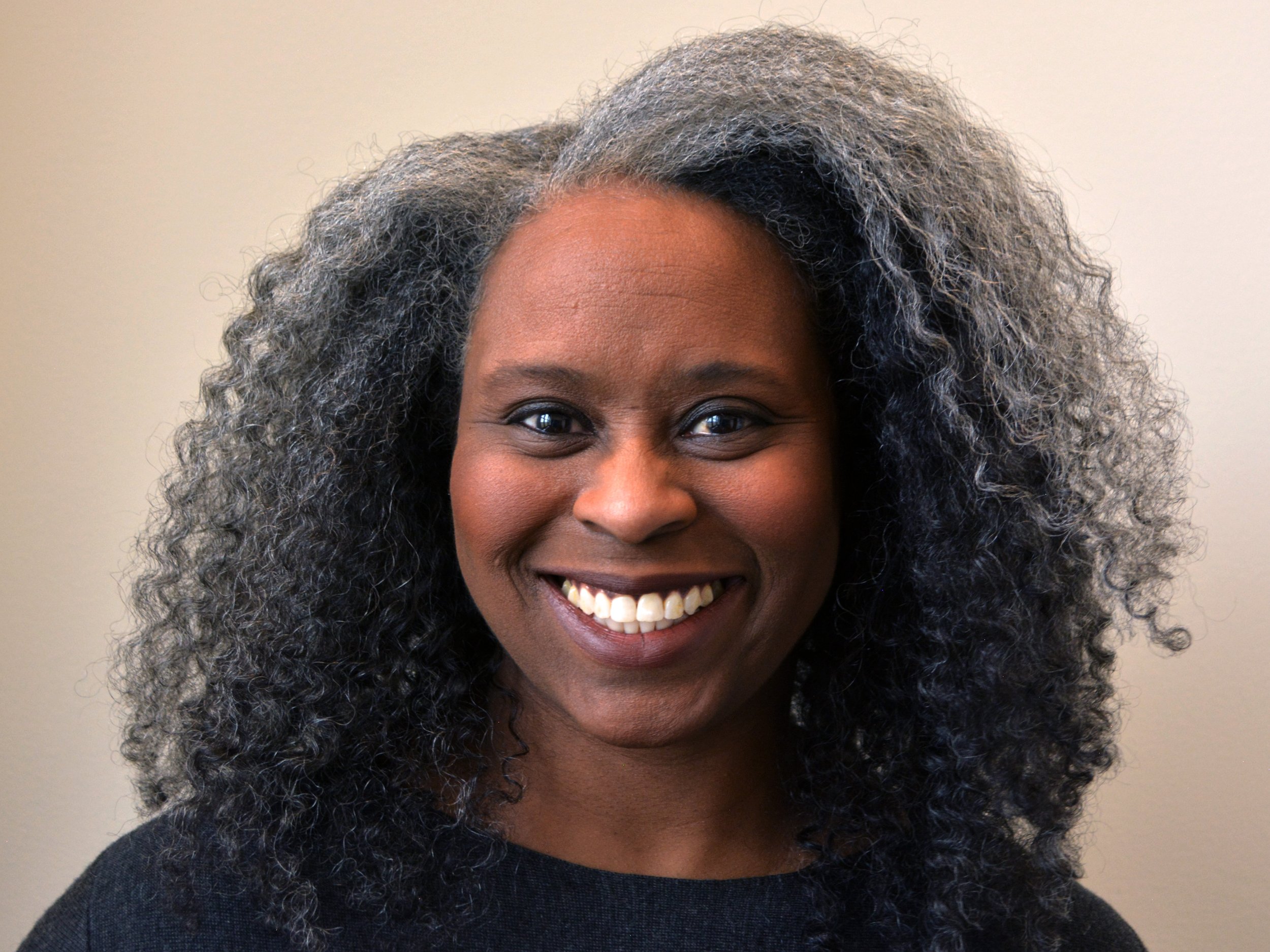 A Black woman with medium brown skin and brown eyes with an open smile. Her hair is salt and pepper gray just below shoulder length naturally coily, curly afro hair. She wears a dark gray felted wool tunic top in front of a light colored background.