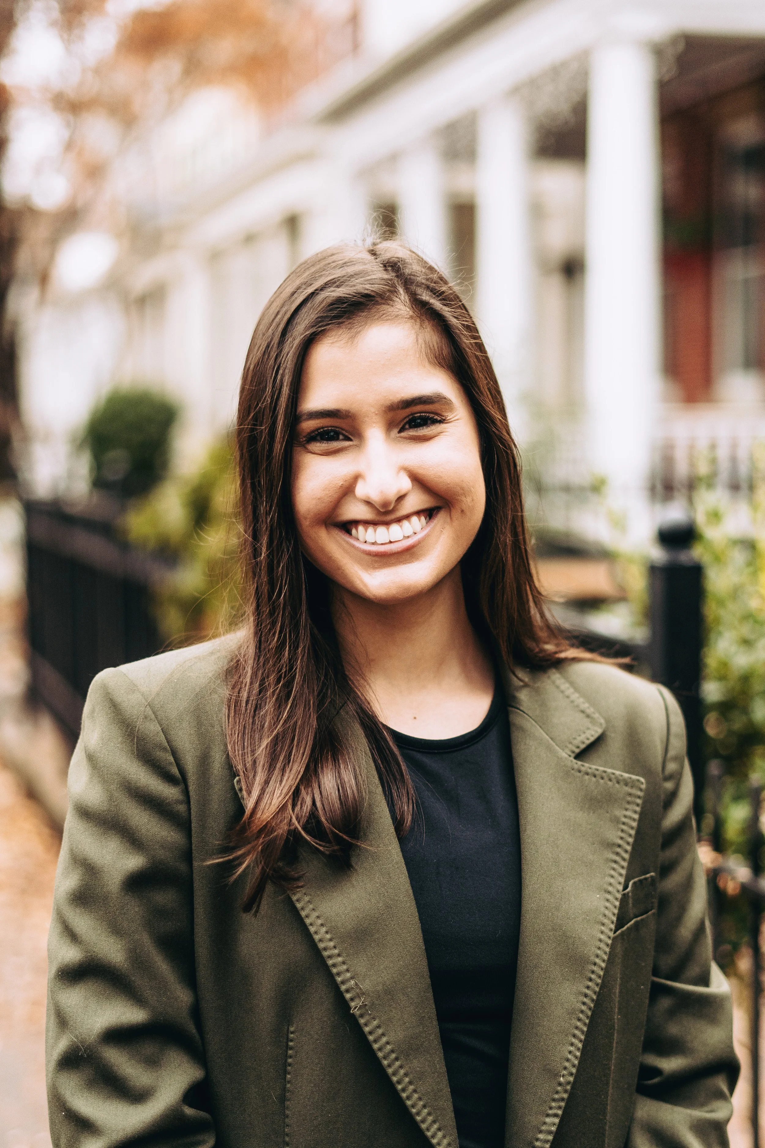 A woman in her 20s with straight, long brown hair and an olive skin tone, wearing a green blazer and black top, smiling at the camera.