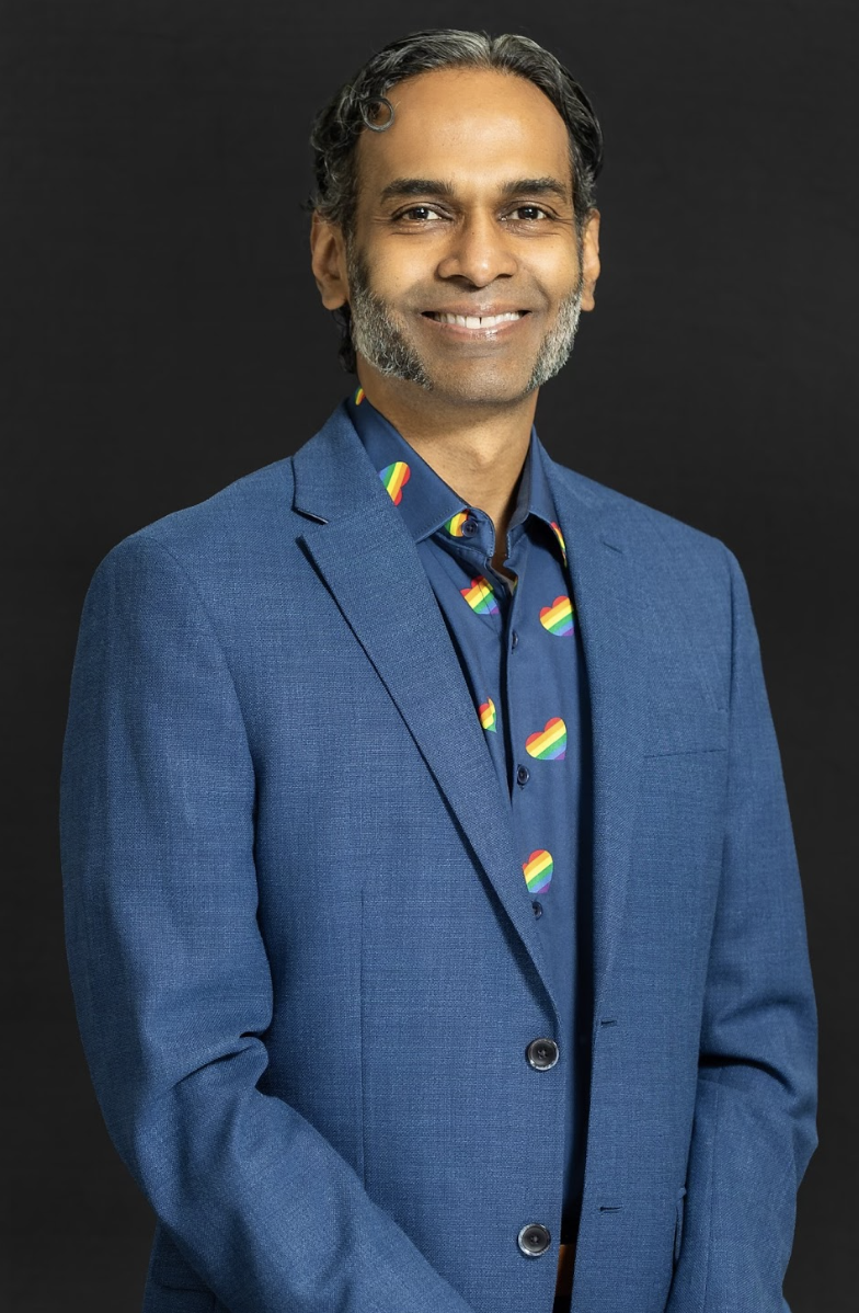 Daniel is smiling at the camera, he is wearing a dark blue shirt with rainbow hearts that signify queer pride
