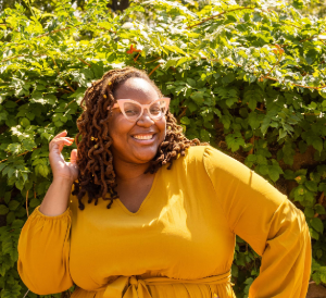 Precious, a brown-skinned Black woman with locs smiles exuberantly at the camera with one hand on her hip and the other bent upward towards her face.
