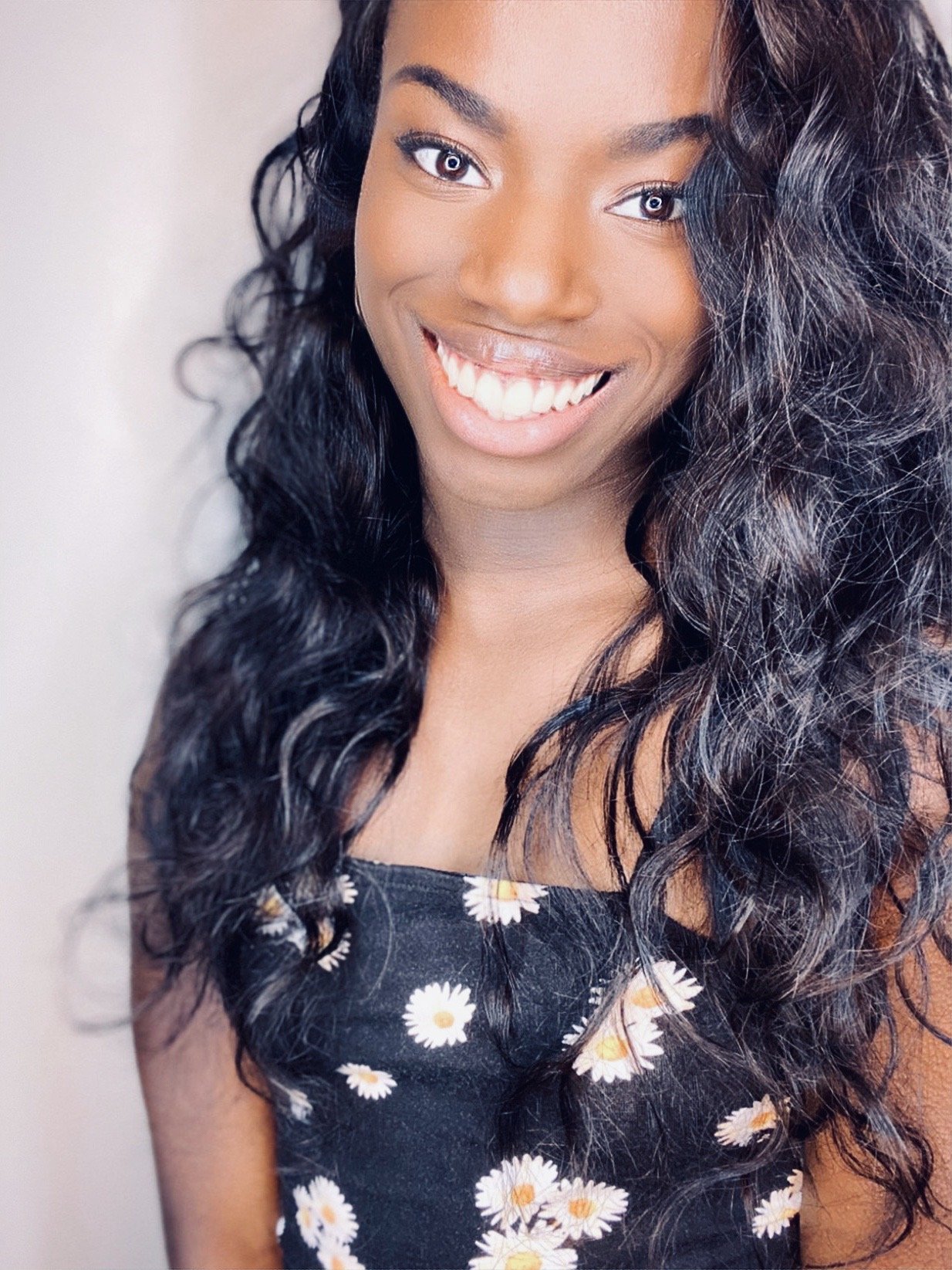 A headshot of Jada, dark skin-tone, smiling directly at the camera wearing a black strap dress with daisies partially covered by loosely curled black hair.