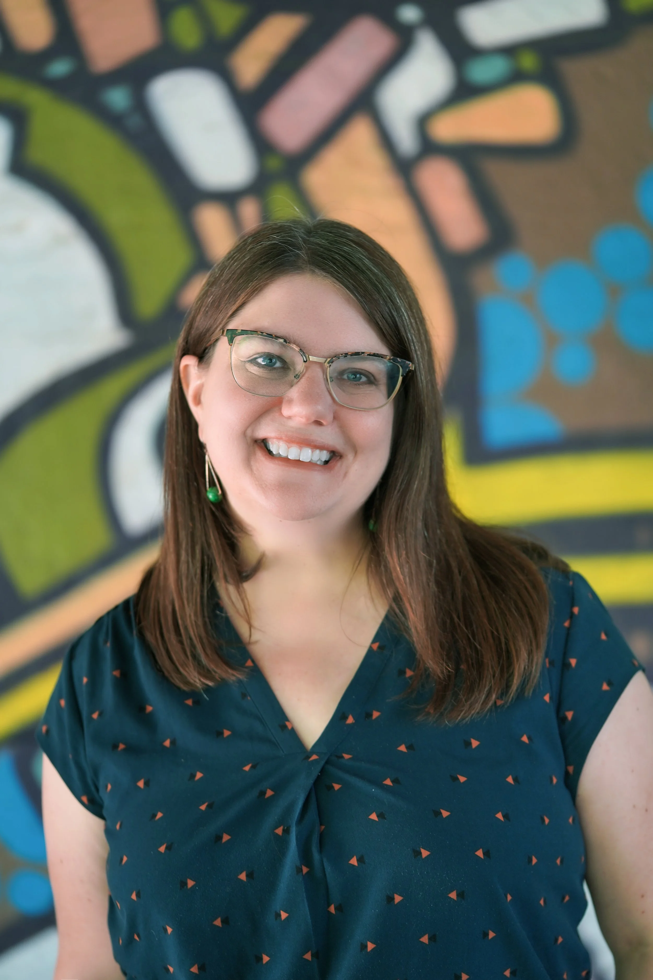 A white woman with long brown hair and glasses, wearing a blue shirt with orange pattern, in front of a colorful mural.