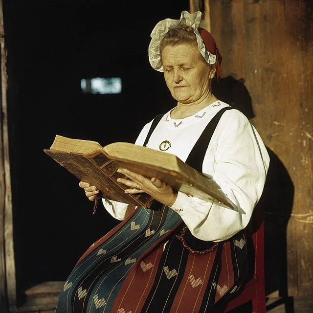 640px-Oude_vrouw_in_klederdracht_voor_houten_huis_die_bijbel_leest,_Bestanddeelnr_254-7433.jpg