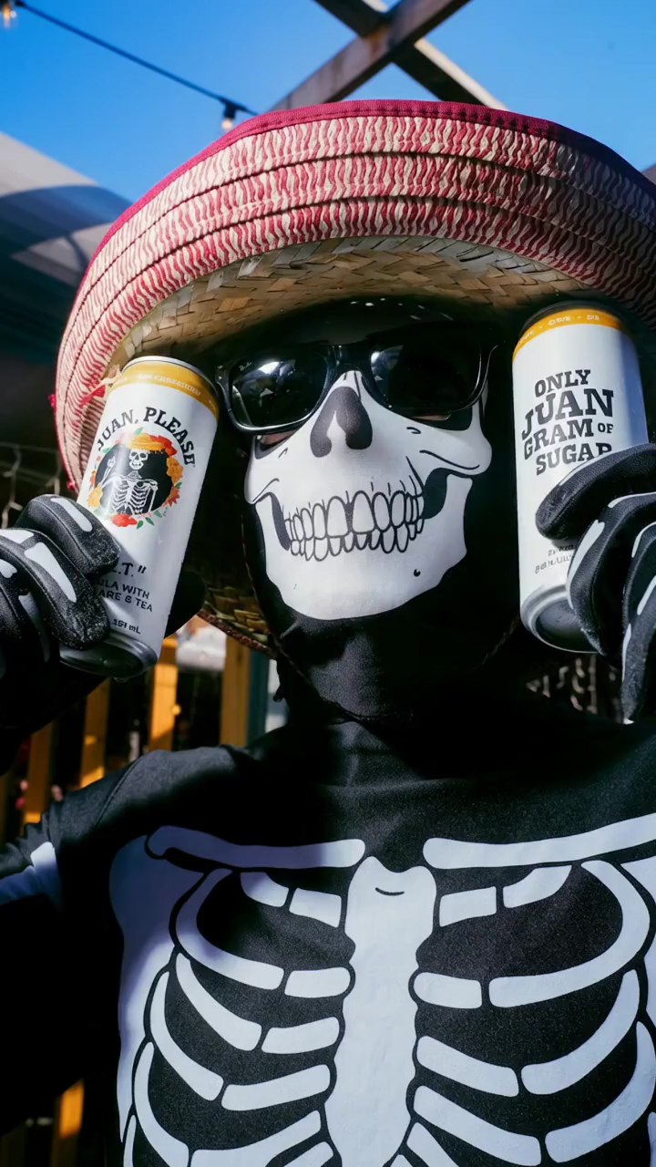A person dressed in a skeleton costume with a skull face mask, wearing sunglasses and a red and white striped sombrero, holding two cans of Juan Good Tea juice, one in each hand, outdoors on a sunny day.
