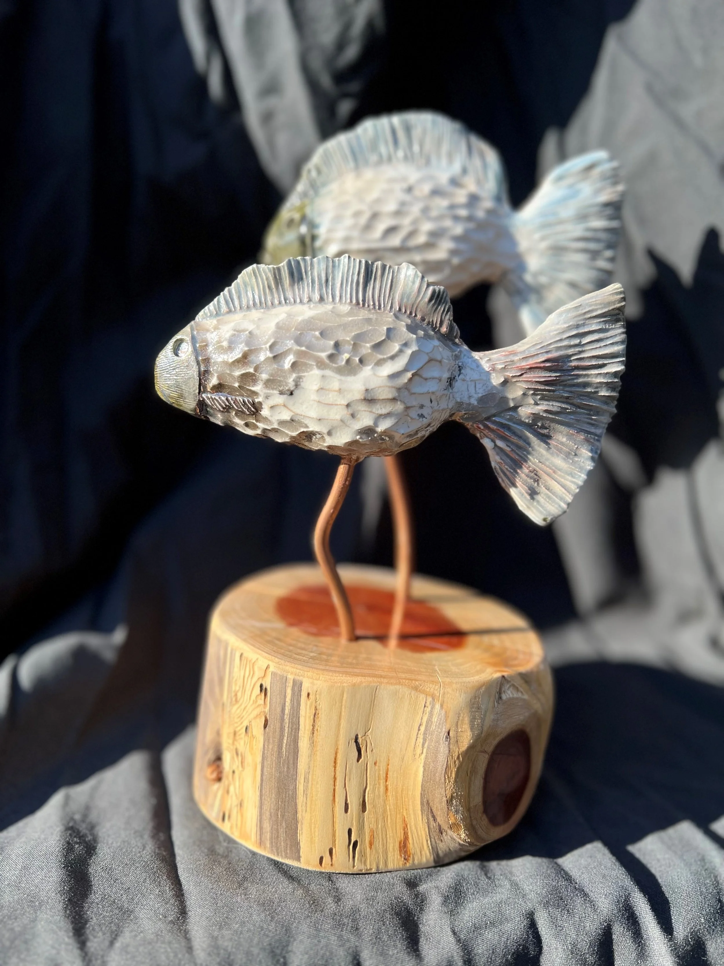 Metal sculpture of a fish with a textured body, mounted on a wooden base with metal legs.