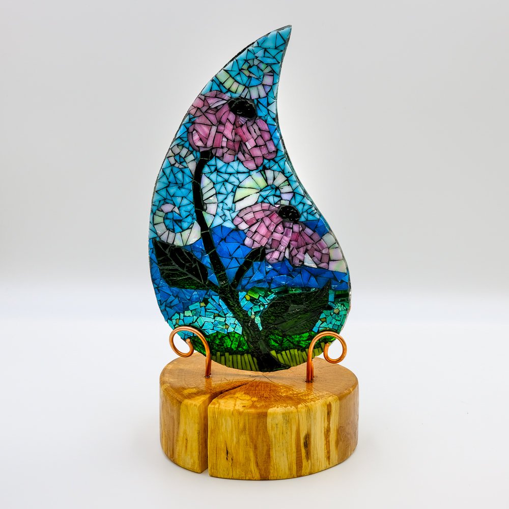 Colorful stained glass sculpture of pink flowers with green leaves and blue sky, mounted on a wooden base.