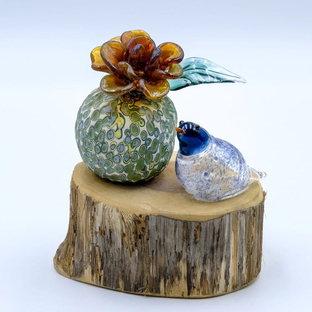 Glass art sculpture of a bird and flower on a wooden base, with a patterned glass flower in a vase and a small glass bird nearby.