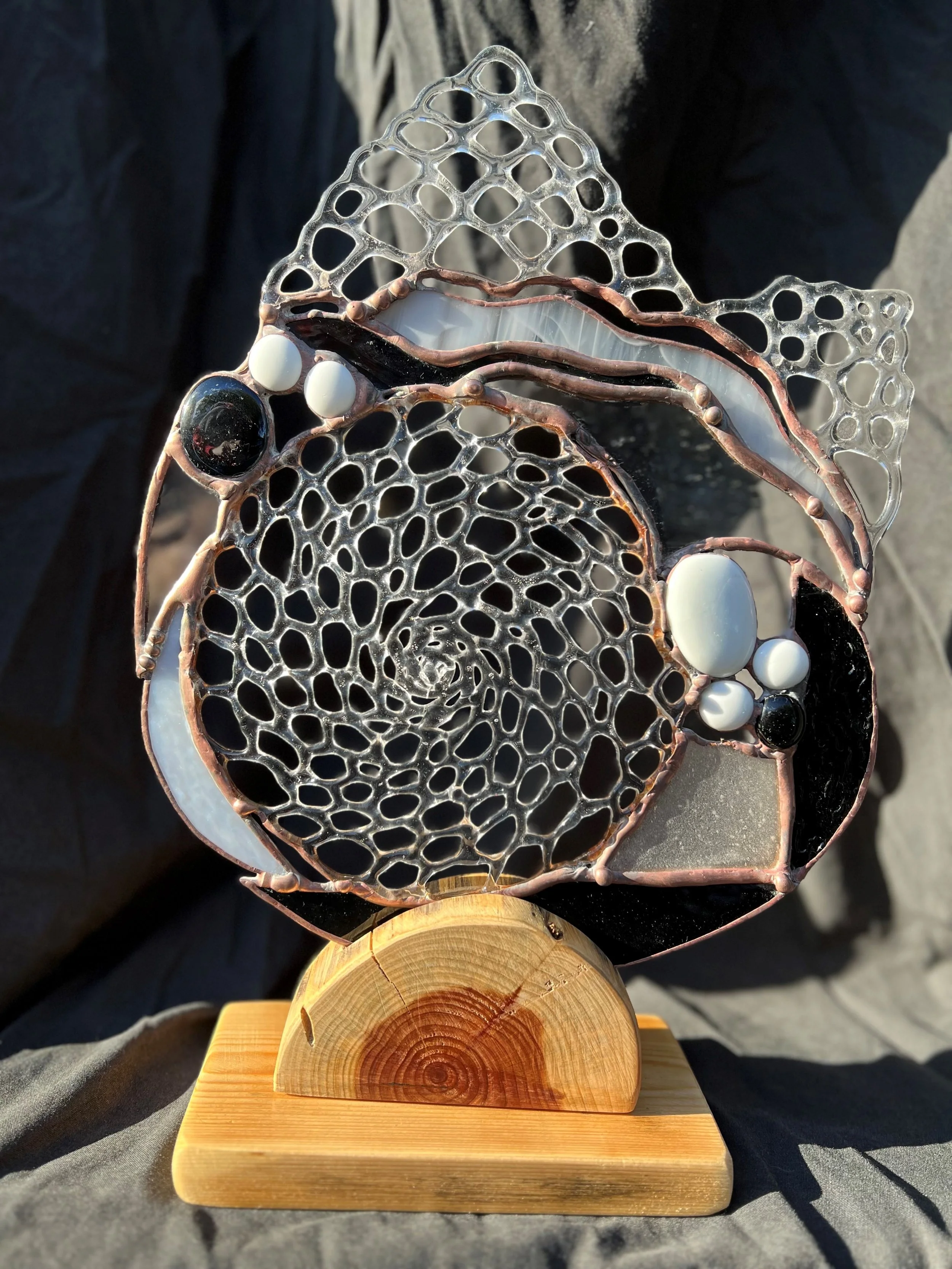 An abstract sculpture resembling a human head with a spiral pattern at the center, made of interconnected metal and glass layers, mounted on a wooden base.