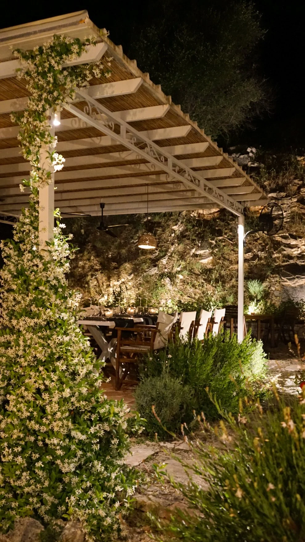 An outdoor dining area at night with a table set for a meal, surrounded by chairs and greenery, under a white pergola with climbing white flowering plants. The background features a rocky hillside with small trees, illuminated by warm lighting.