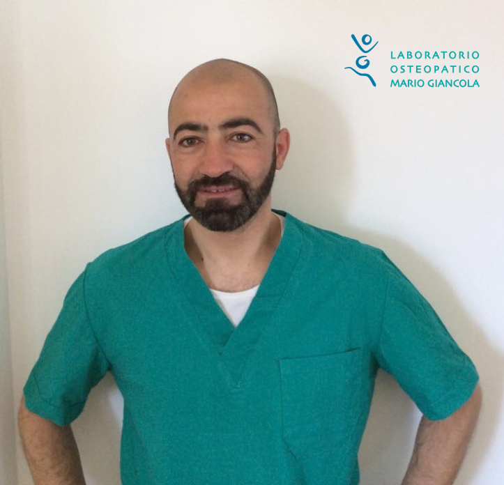 Man in teal medical scrubs standing with hands on hips, smiling, in front of a white wall with a logo for Laboratorio Osteopatico Mario Giancola.