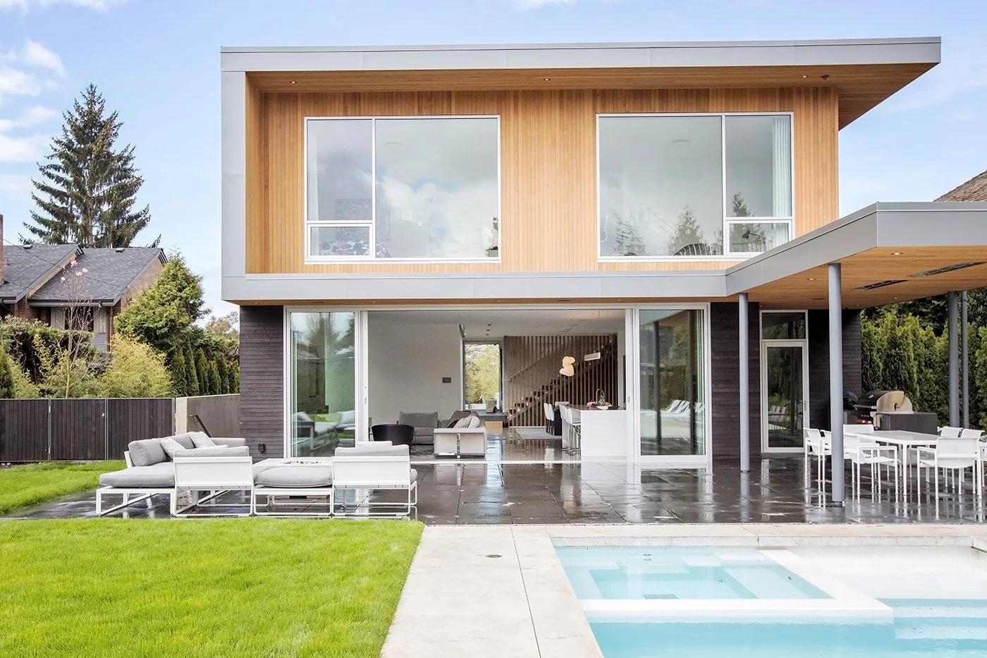 Modern two-story house with large glass windows, wooden and dark exterior finishes, outdoor patio with seating and dining area, lush green lawn, and a swimming pool in the backyard.