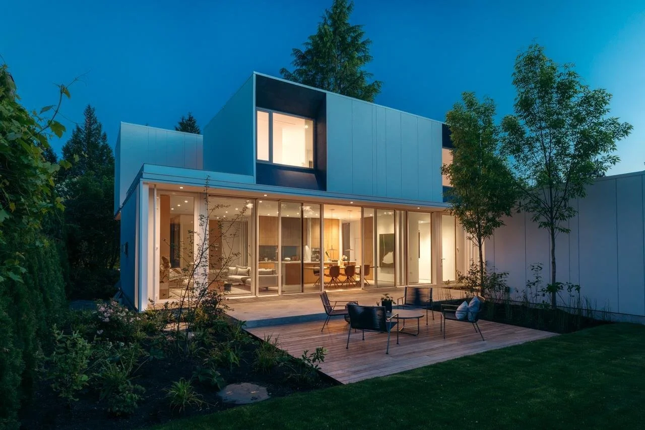 Modern two-story house with large glass doors opening to a wooden patio, outdoor seating, and surrounding greenery in the evening.