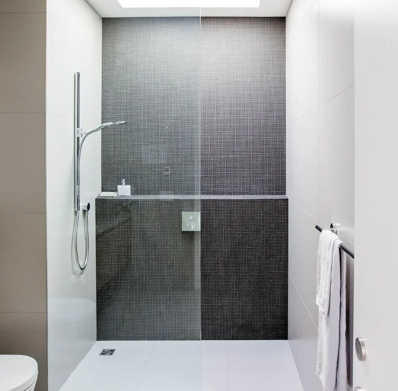 Modern shower area with gray mosaic tile wall, white and beige wall tiles, and a glass divider, including a showerhead, soap dispenser, and towel bar.