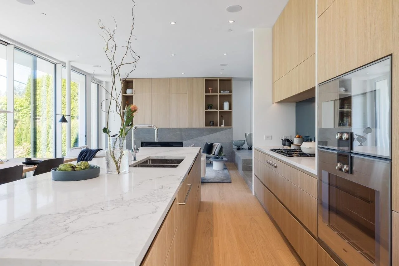 Modern kitchen with white marble island, light wood cabinets, built-in oven, and open living area with large windows and green view